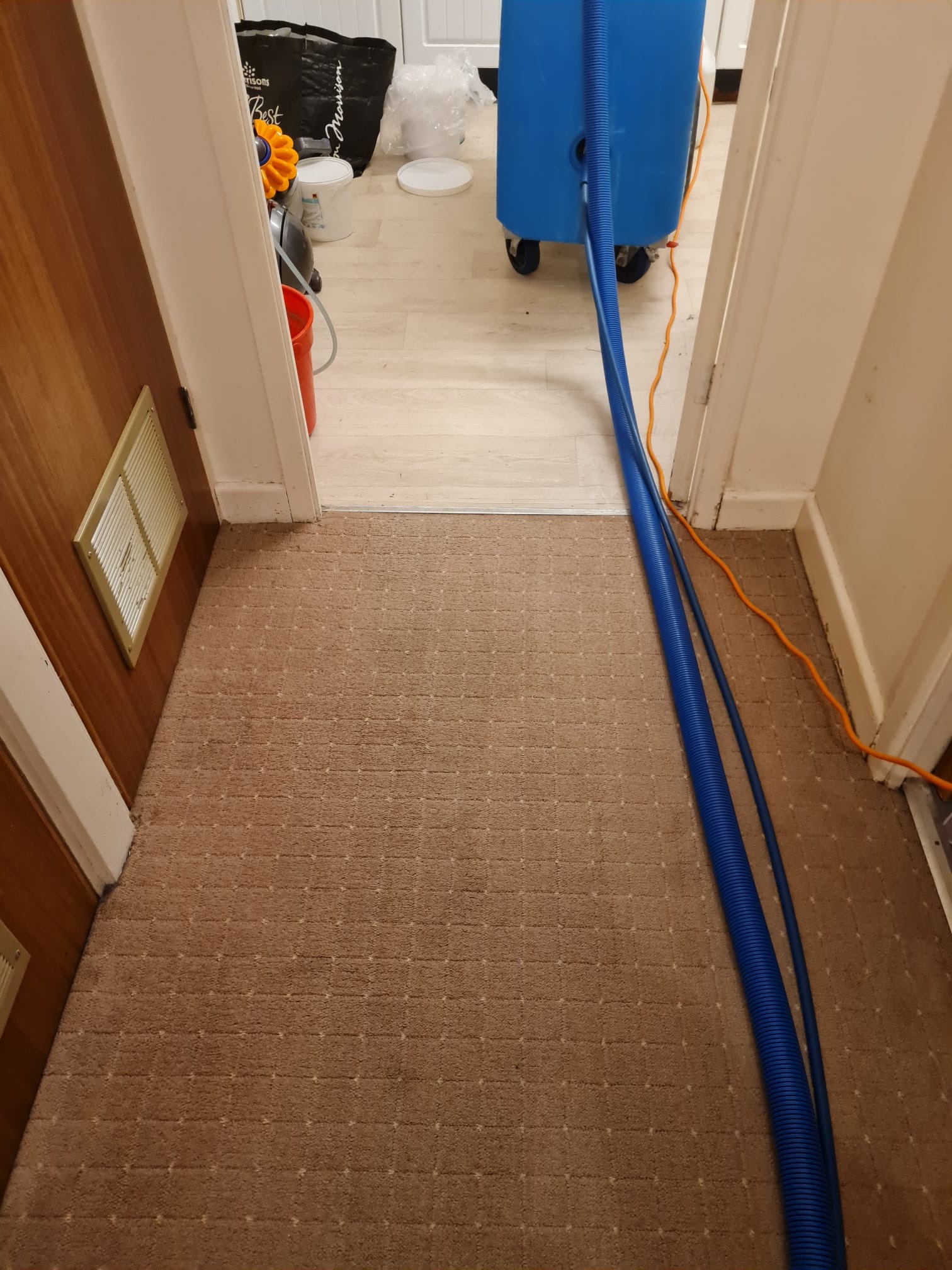 Brązowy dywan z widocznym wężem odkurzacza piorącego podczas pracy, z boku kratka wentylacyjna w ścianie.