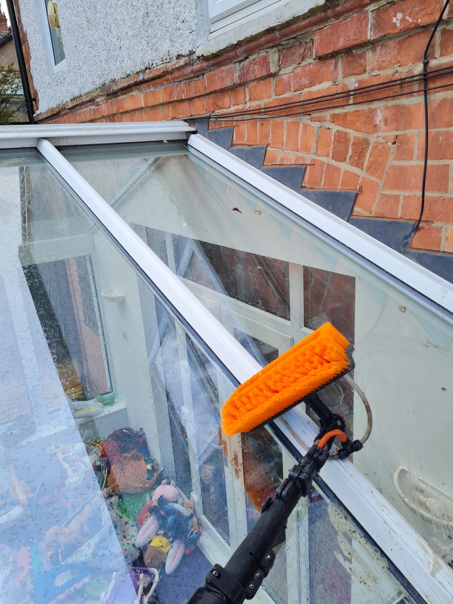 Mycie przeszklonego dachu oranżerii za pomocą szczotki na teleskopowym wysięgniku, widoczne krople wody i refleksy na szybach, w tle ceglana ściana budynku.