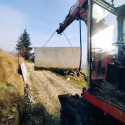 BUDUJEMY Z PASJĄ Krzysztof Pastwa - Ciężki, betonowy element konstrukcyjny podnoszony przez dźwig na nierównym, wiejskim terenie z widocznym lasem w tle, widziany z perspektywy kabiny operatora dźwigu.