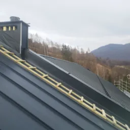Ciemnoszary dach z blachy na tle zalesionych wzg&oacute;rz, widoczny komin z izolacją i drewniane rusztowanie dachowe.