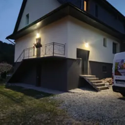 Dwukondygnacyjny dom z balkonem, oświetlony zewnętrznymi lampami, z częściowo widocznym samochodem dostawczym z grafiką związaną z dachem. Zakamarki w wieczornym świetle.