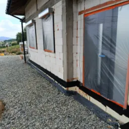 Ściana domu w trakcie budowy z oknami zabezpieczonymi folią, widoczne bloczki z betonu kom&oacute;rkowego, izolacja fundament&oacute;w i podsypka żwirowa wok&oacute;ł budynku.