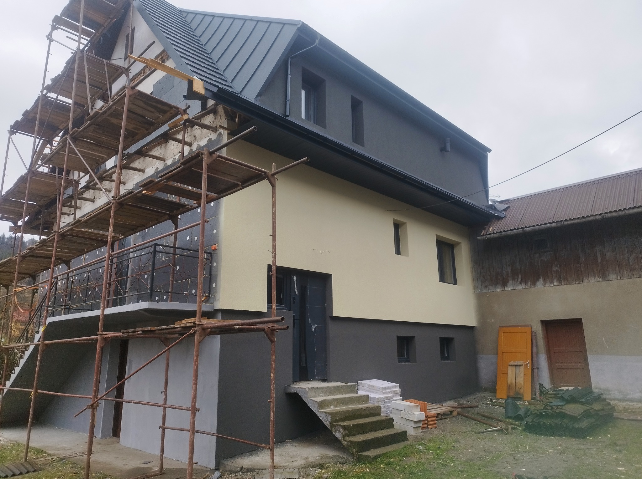 Dom w trakcie ocieplania elewacji z widocznymi rusztowaniami, częściowo otynkowany, z nowym dachem i stosem płyt styropianowych na podwórku.