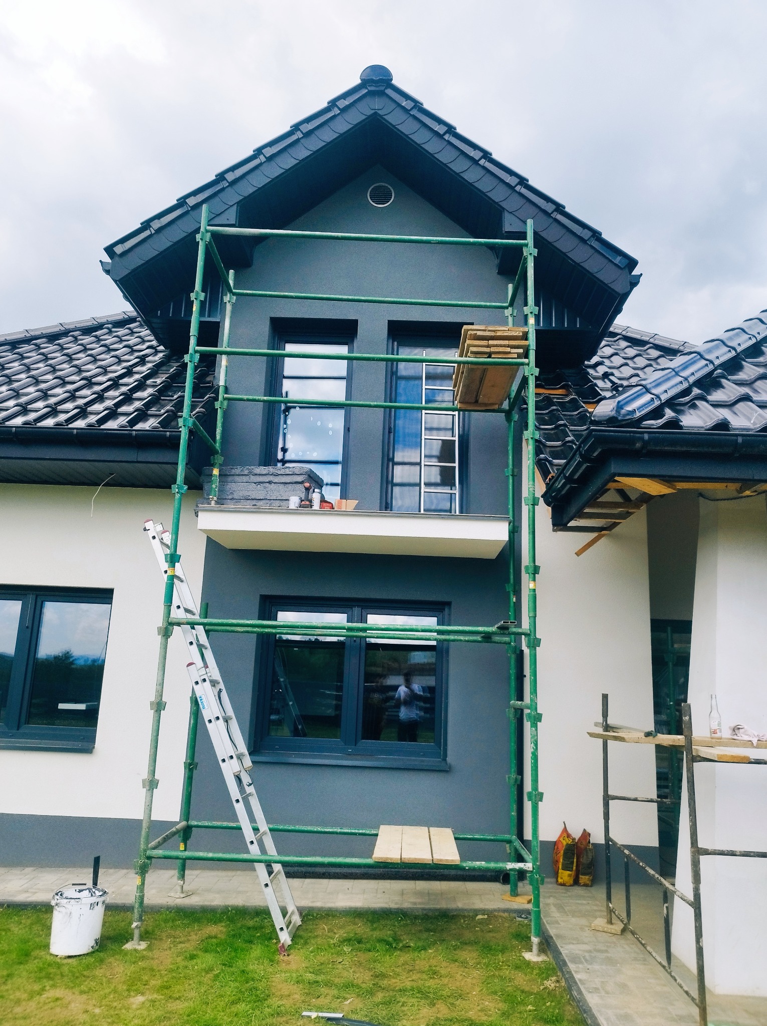 Dwukondygnacyjny dom w trakcie prac elewacyjnych, z widocznym rusztowaniem, drabiną opartą o ścianę i częściowo pomalowaną elewacją na szaro