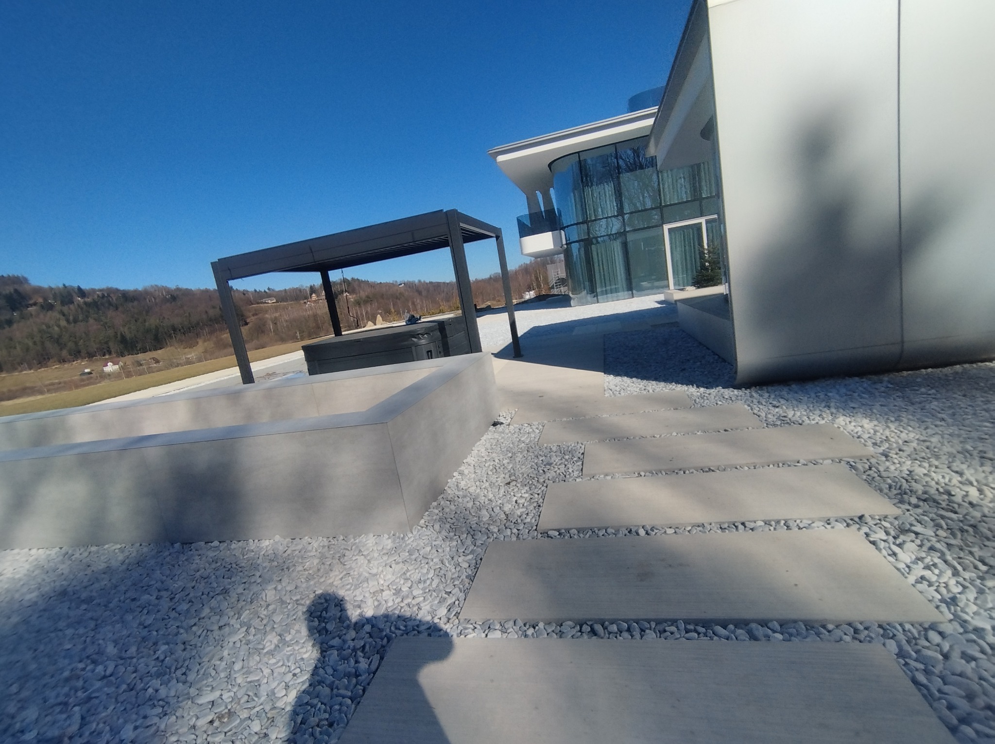 Nowoczesny dom z dużymi przeszkleniami i tarasem z pergolą w Sidzinie, wykończony białym kamieniem ozdobnym. Widoczne betonowe płyty chodnikowe.