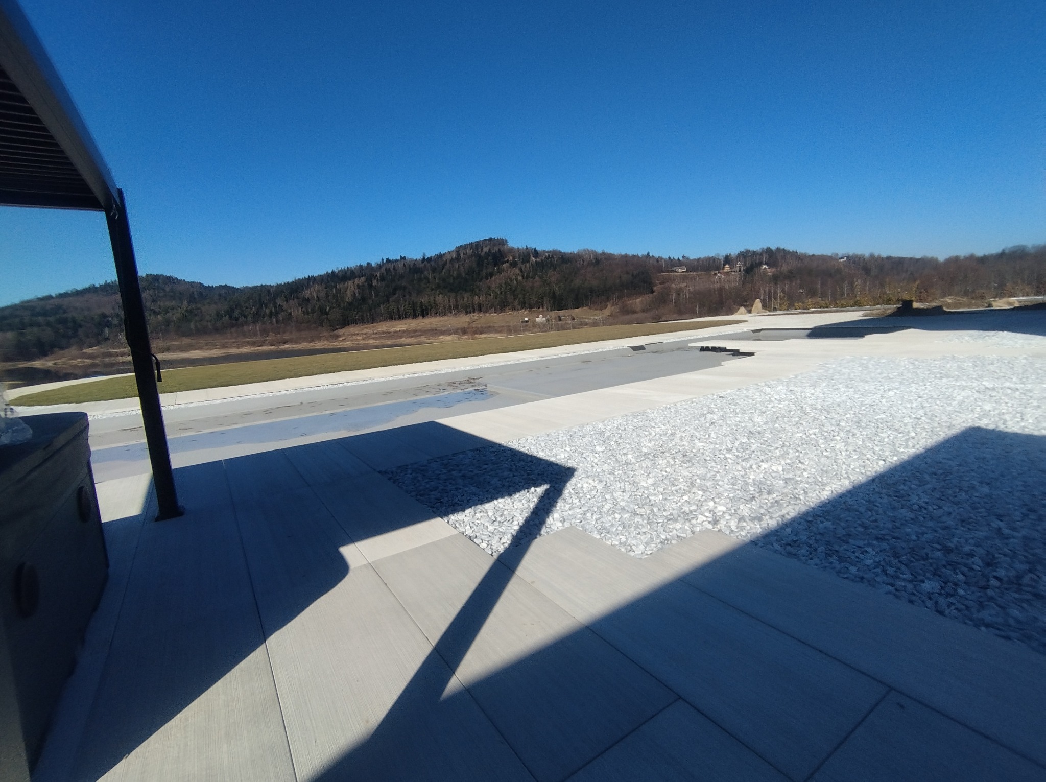 Nowoczesny taras wyłożony szarymi płytami z elementami dekoracyjnymi z białego żwiru, widok na zalesione wzgórza w tle.