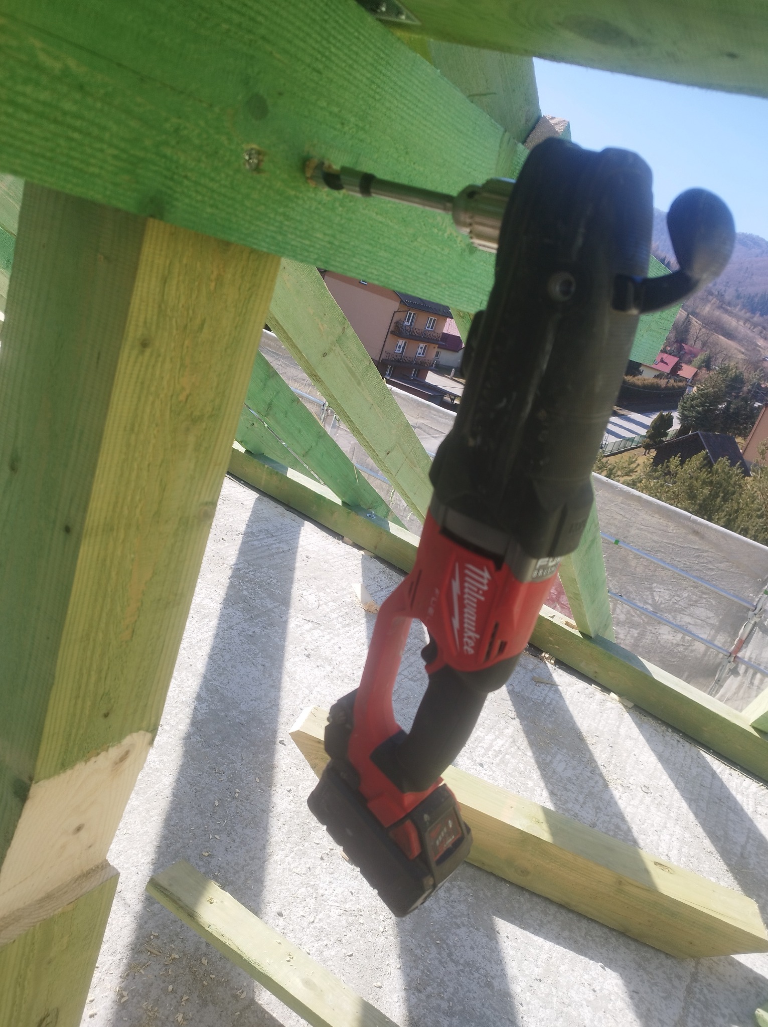 Wiertarka Milwaukee wkręcająca śrubę w zieloną drewnianą konstrukcję dachu na tle gór i budynków.