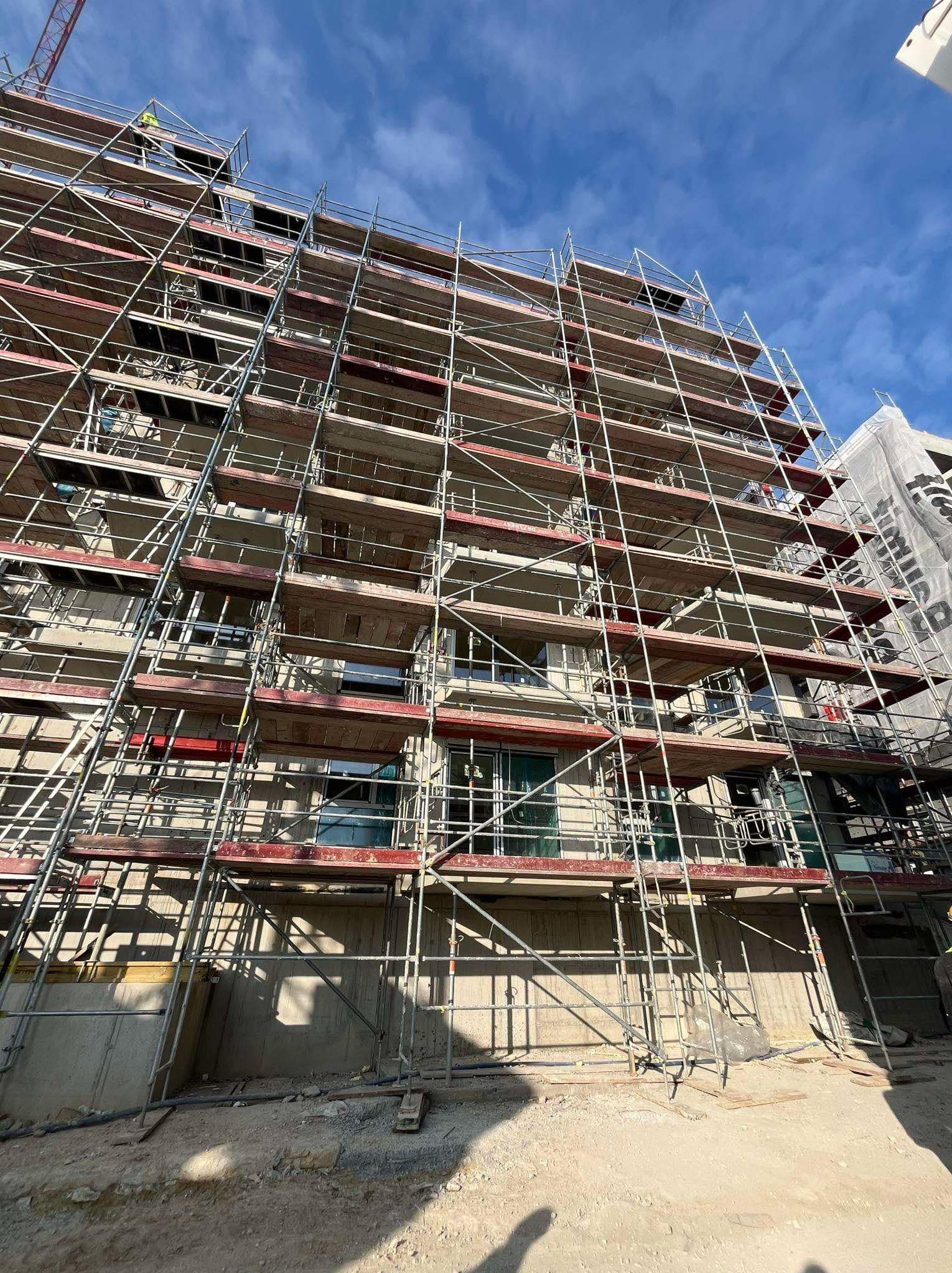 Wielopiętrowy budynek w trakcie budowy, z widocznymi rusztowaniami, surową elewacją i niebieskim niebem w tle.
