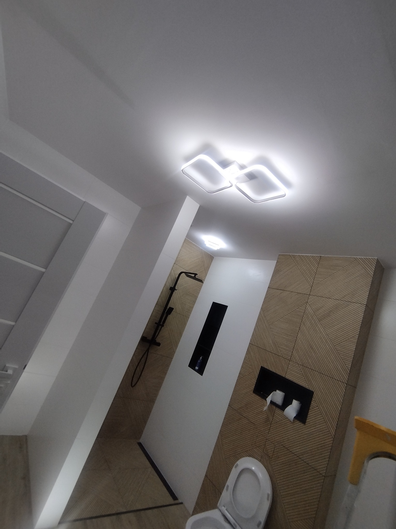 Nowoczesna łazienka z geometrycznymi płytkami na ścianie, oświetleniem LED i czarną armaturą prysznicową.