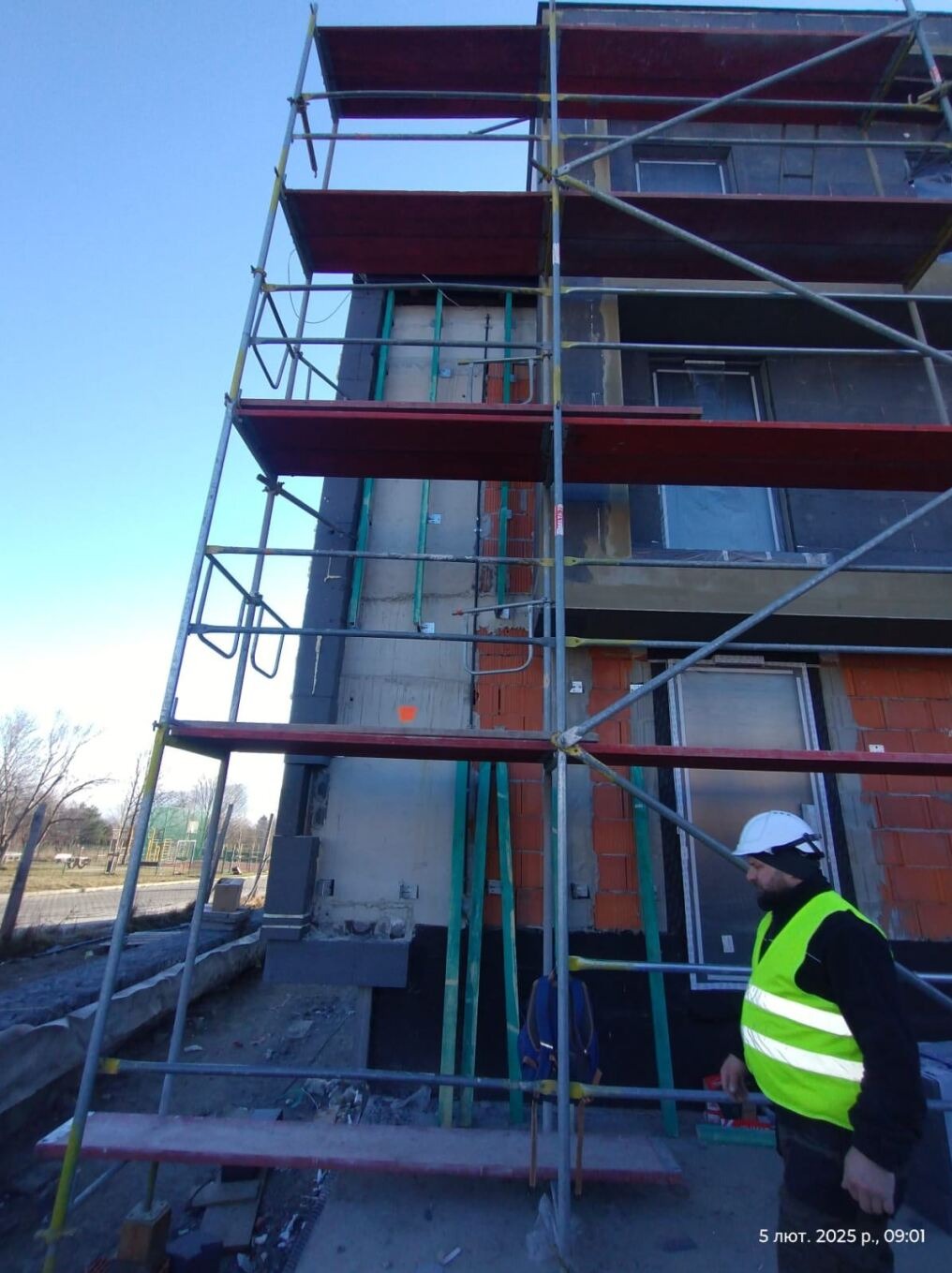 Budowa wielopiętrowego budynku z cegły, widok z rusztowania, pracownik w kasku i kamizelce odblaskowej kontroluje postęp prac, widoczne okna i instalacje.