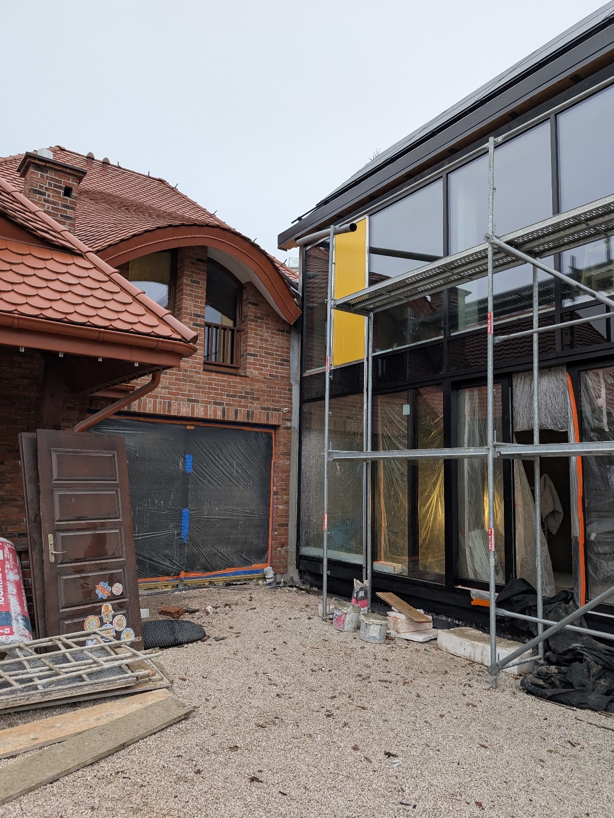 Budynek w trakcie budowy: część murowana z czerwoną dachówką i część nowoczesna z dużymi oknami, rusztowania, materiały budowlane na ziemi.