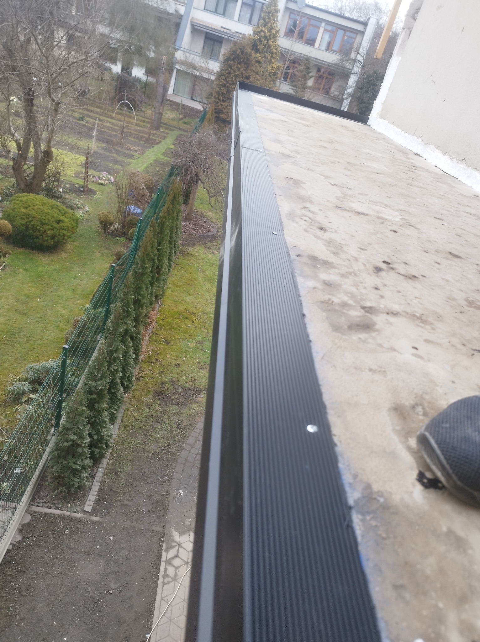 Widok z góry na płaski dach z zamontowaną, ciemną obróbką blacharską, w tle ogród z tujami i budynki mieszkalne.