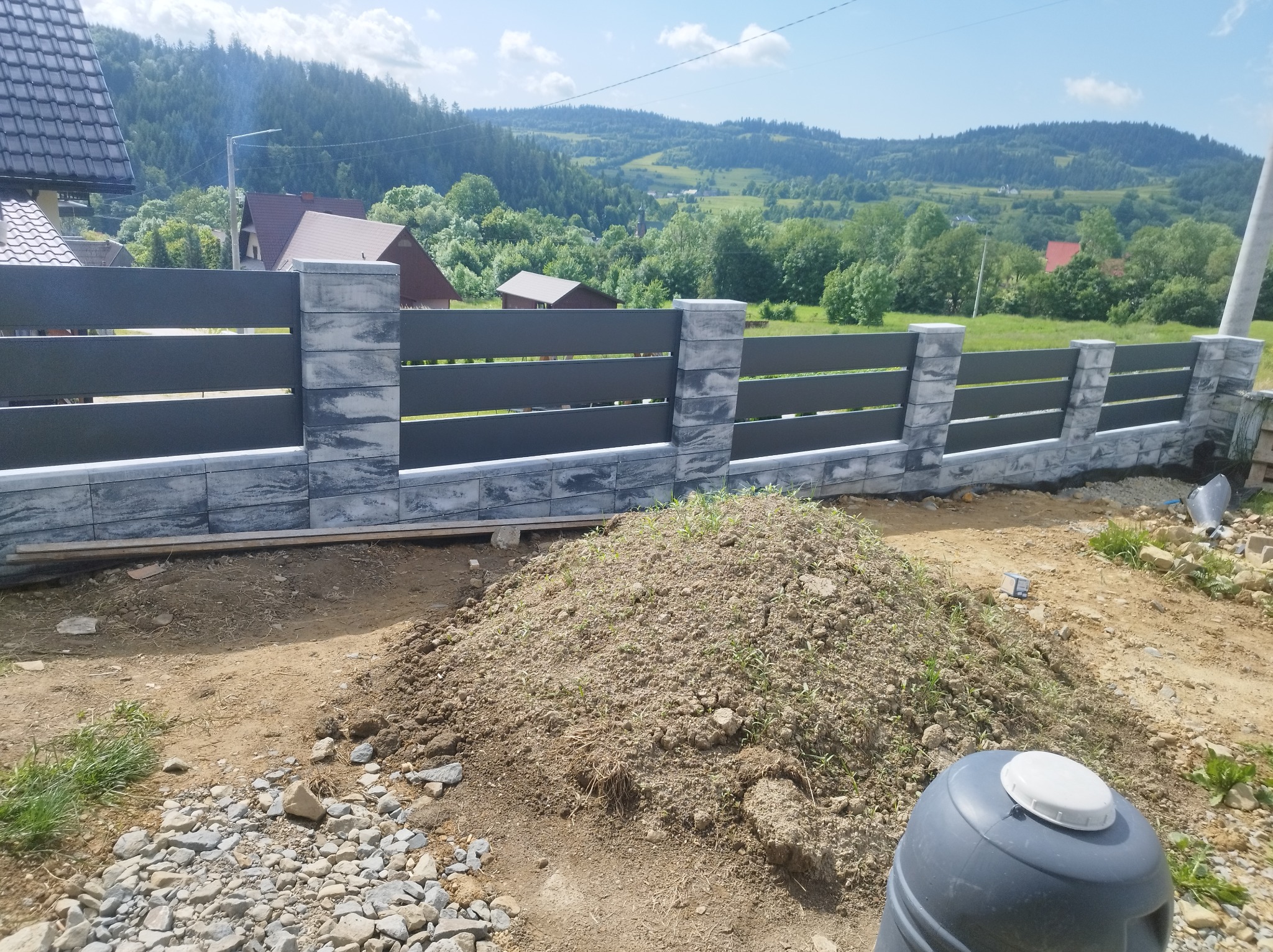 Nowoczesne ogrodzenie panelowe z szarymi, betonowymi słupkami na tle malowniczego krajobrazu górskiego w słoneczny dzień. Widoczna sterta ziemi i elementy budowlane.