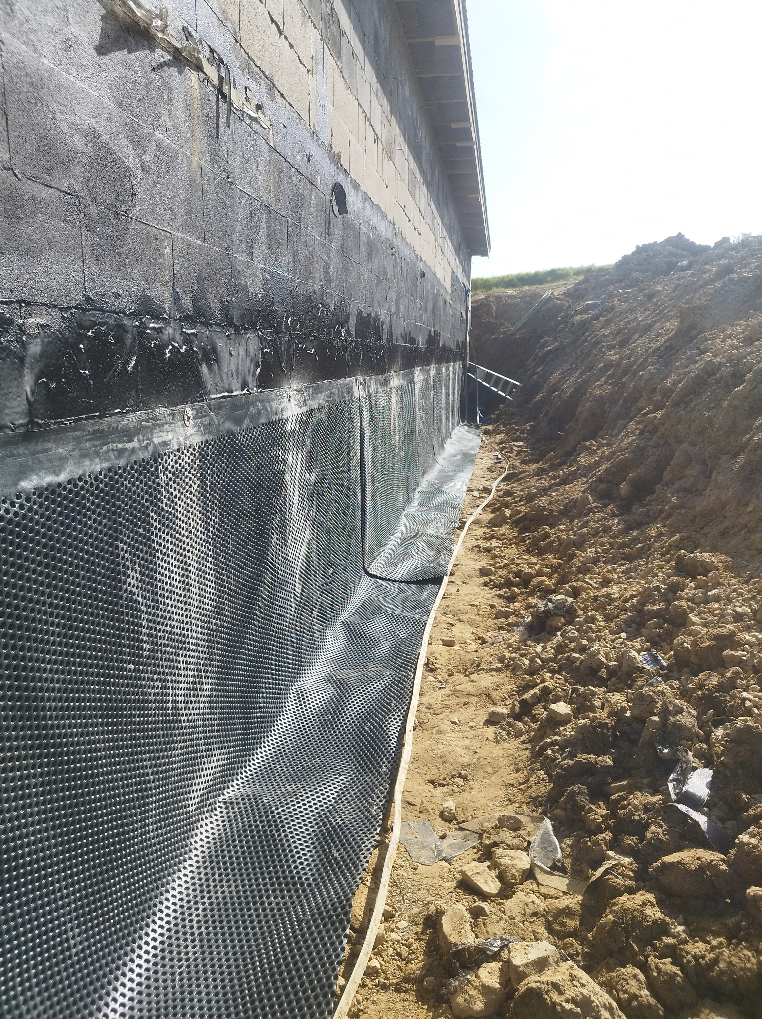 Izolacja pionowa fundamentu budynku z bloczków betonowych, zabezpieczona folią kubełkową i masą bitumiczną, widoczny wykop ziemny.