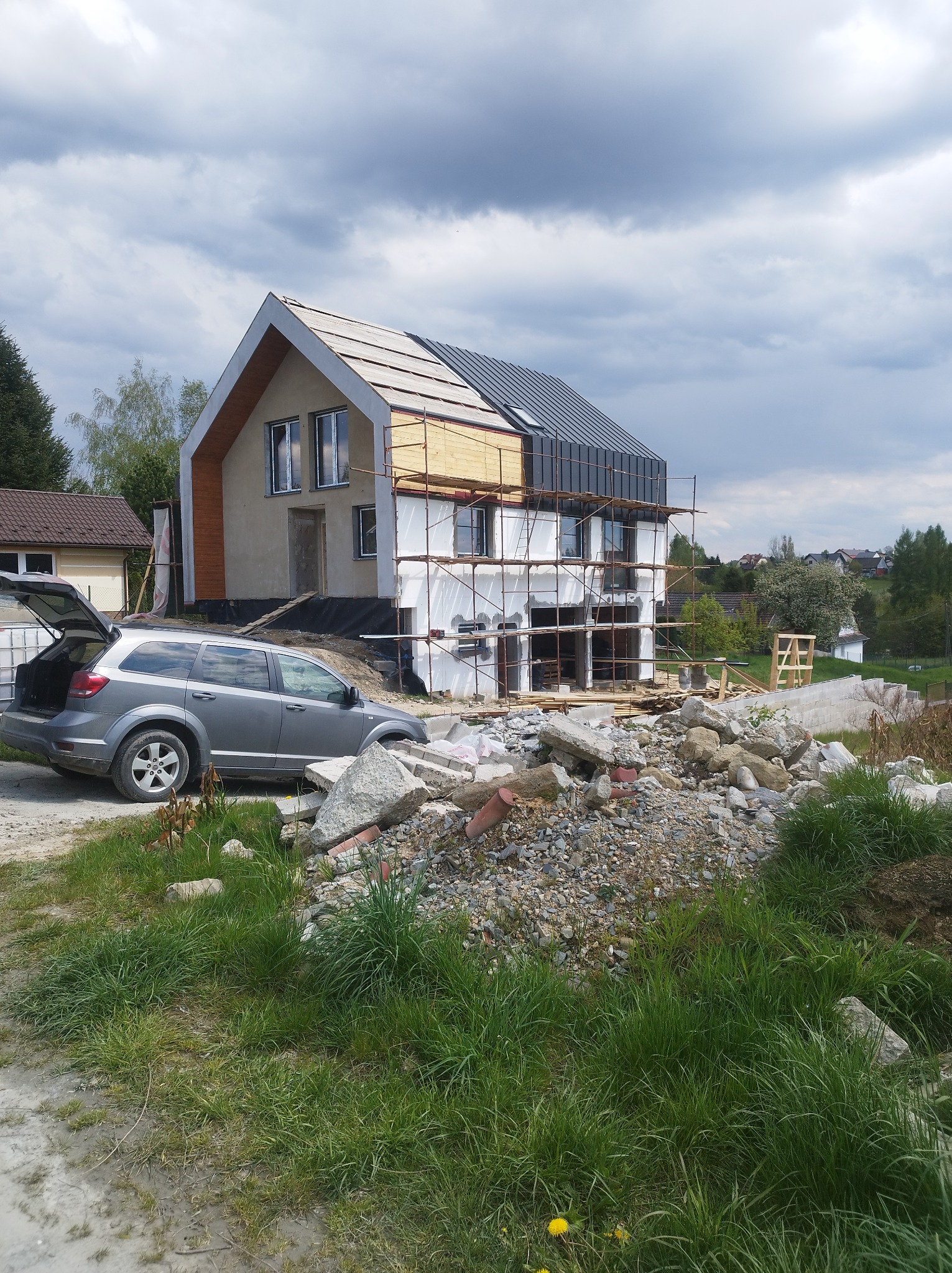 Budowa domu murowanego z częściowo ukończonym dachem z blachy na rąbek stojący, widoczne rusztowania i samochód z otwartym bagażnikiem na tle zachmurzonego nieba.