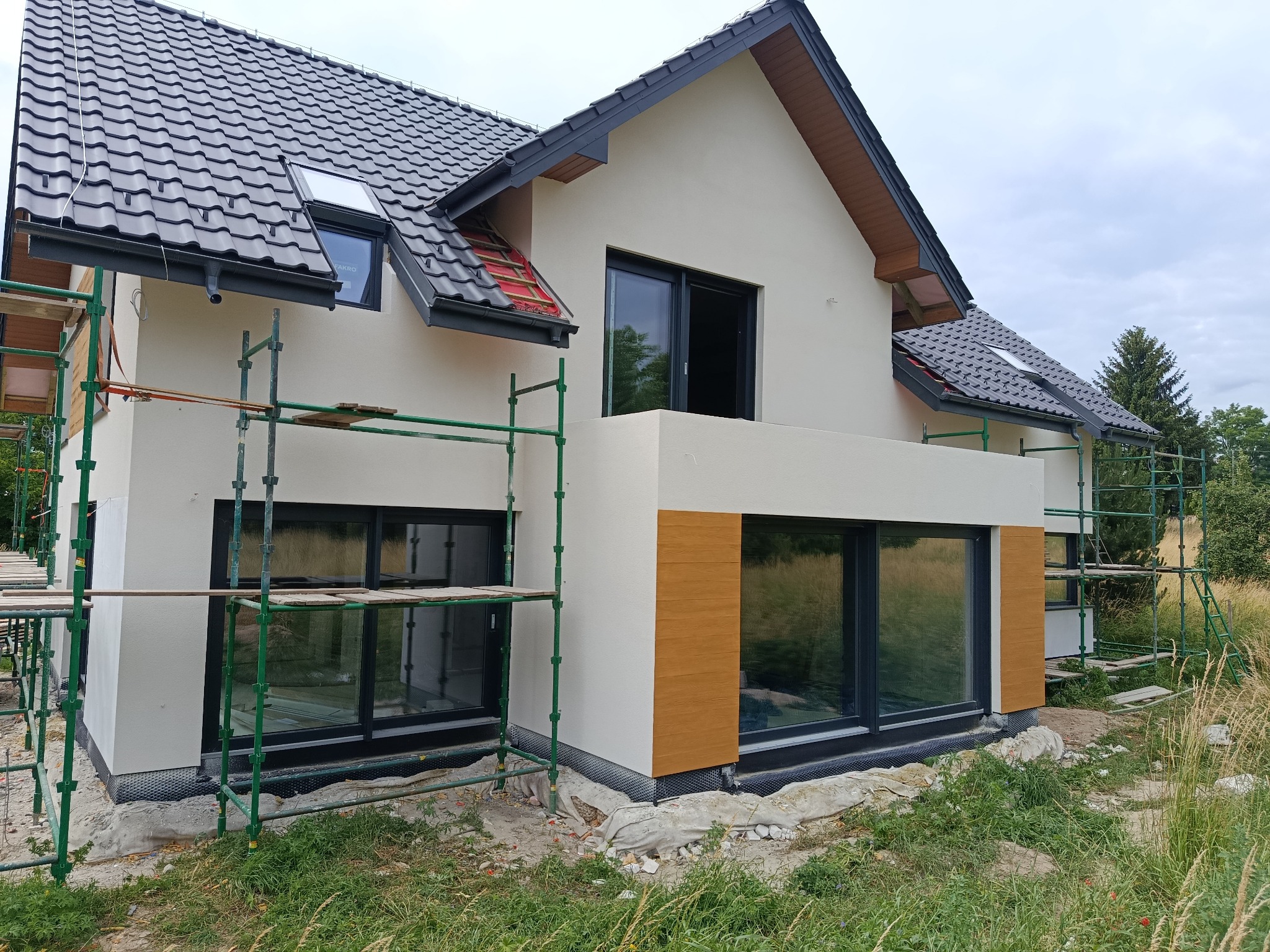 Nowoczesny dom w trakcie wykończenia elewacji, z widocznymi rusztowaniami i fragmentami nieotynkowanej powierzchni. Dach z ciemnej dachówki.
