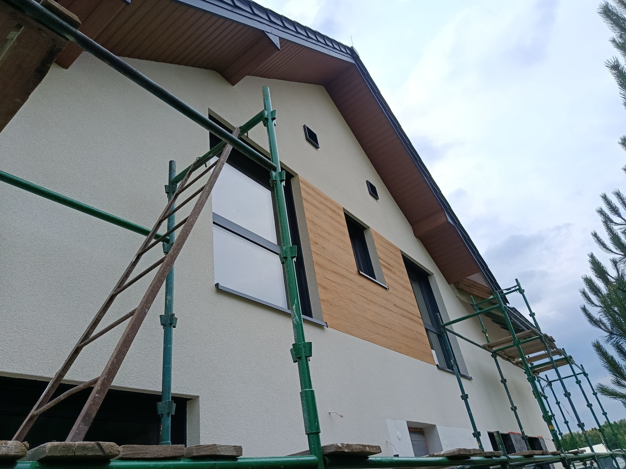 Elewacja domu z fragmentem drewnianej okładziny, widoczna z poziomu rusztowania. Ujęcie z dołu, niebo w tle.