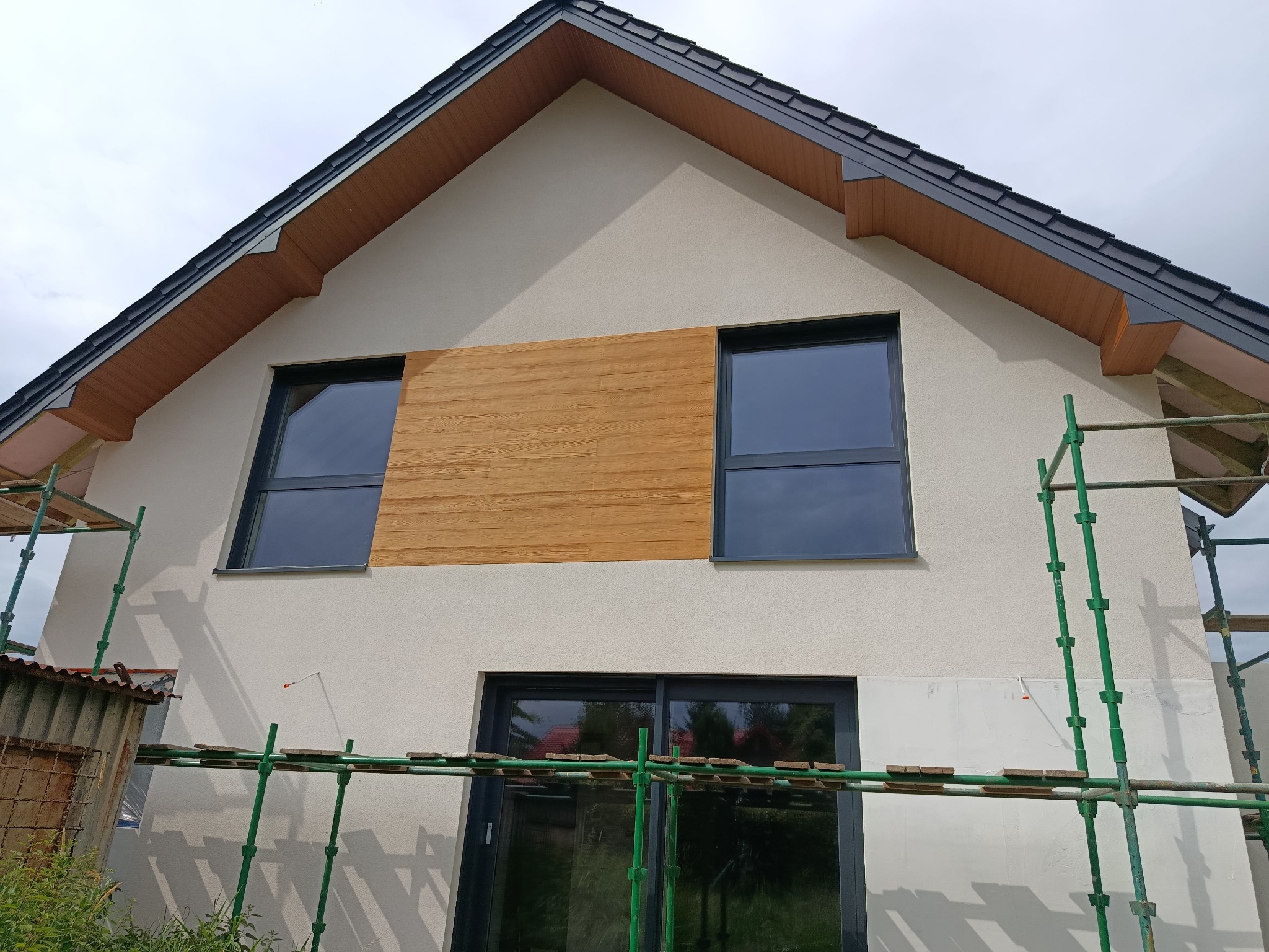 Elewacja domu jednorodzinnego z oknami i drewnianym panelem dekoracyjnym, widok z rusztowania w Sidzinie. Kremowa elewacja, grafitowy dach.