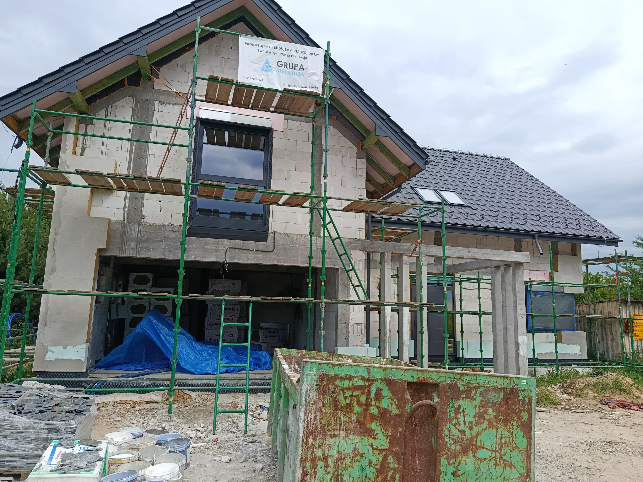 Dom w stanie surowym otwartym, z rusztowaniem i kontenerem na gruz. Widoczne elementy dachu i okna. Na rusztowaniu baner firmy budowlanej.