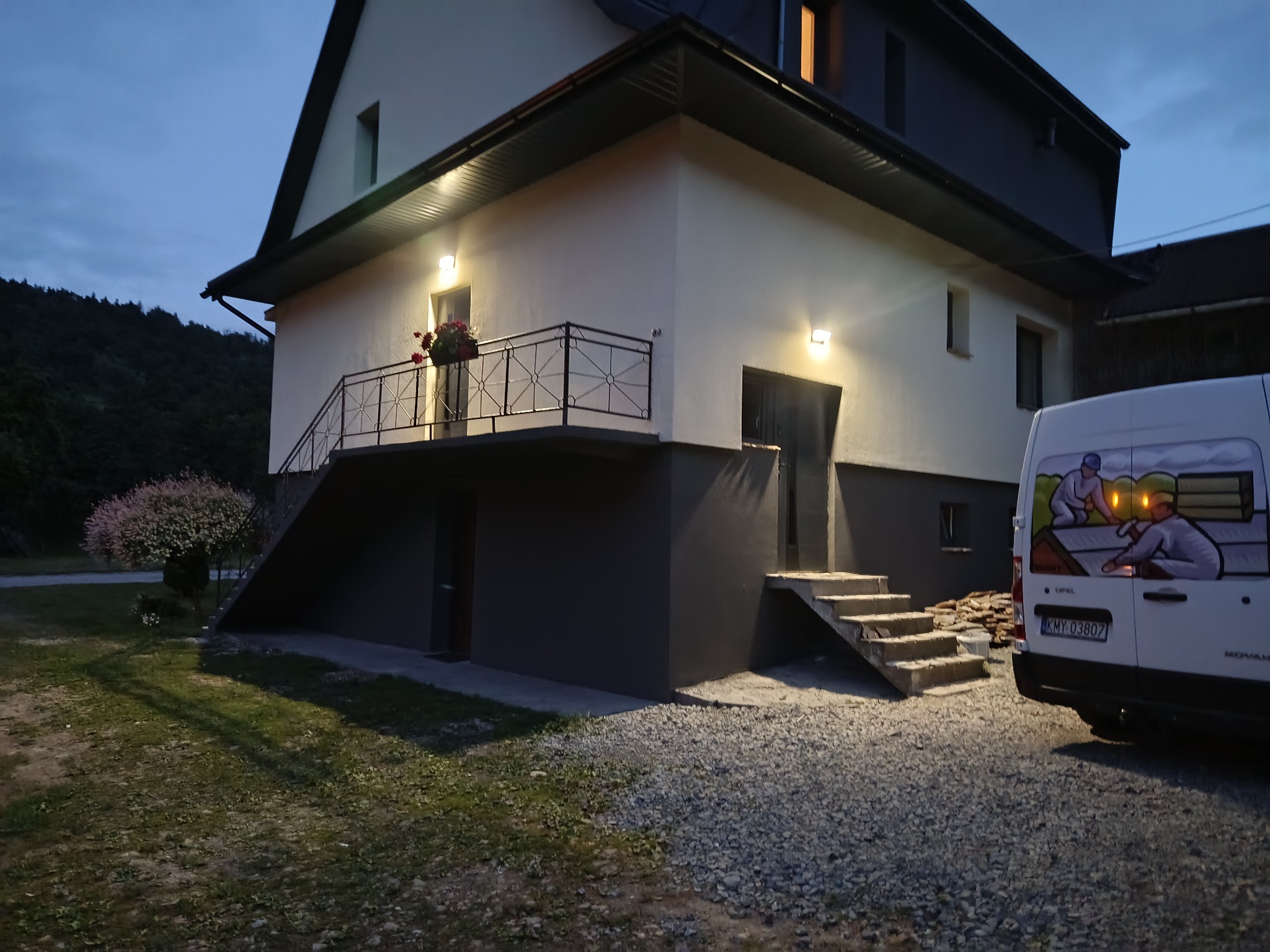 Dwukondygnacyjny dom z balkonem, oświetlony zewnętrznymi lampami, z częściowo widocznym samochodem dostawczym z grafiką związaną z dachem. Zakamarki w wieczornym świetle.