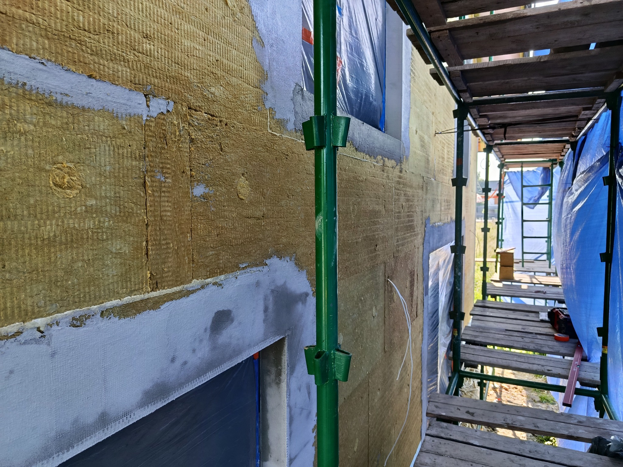 Ocieplanie ściany budynku wełną mineralną, widoczne rusztowanie i okna zabezpieczone folią. Etap prac przed nałożeniem tynku. Detal mocowania rusztowania.