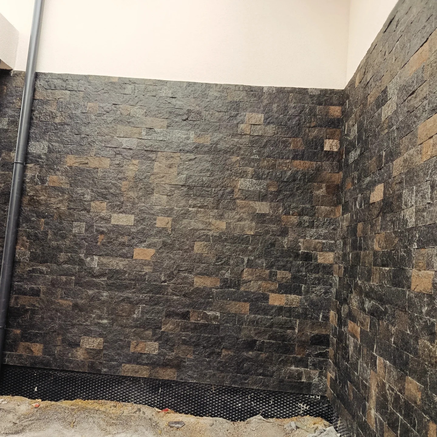 Ściana wykończona dekoracyjną, ciemną okładziną kamienną z fragmentem instalacji i podłoża w dolnej części kadru. Ujęcie z perspektywy narożnika.
