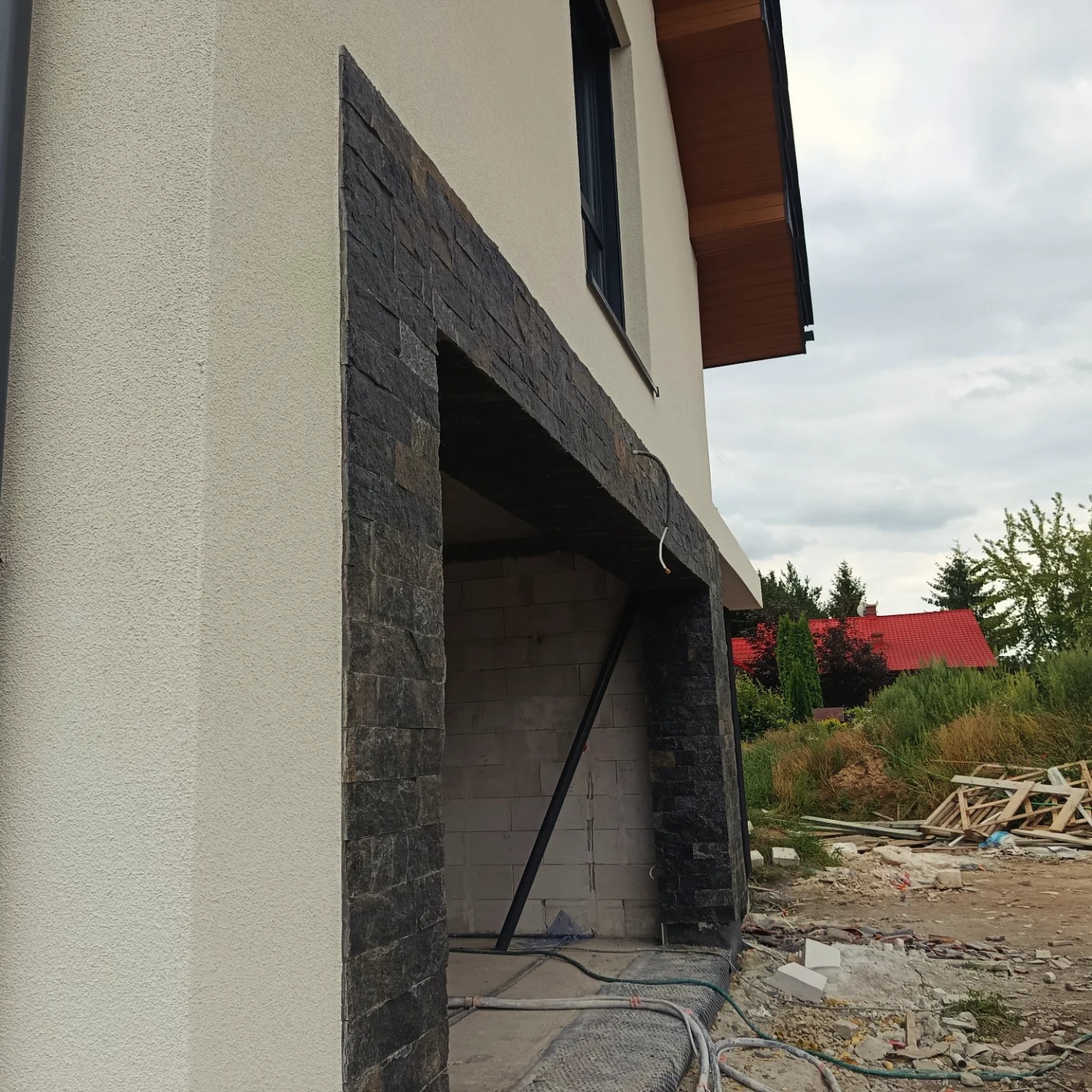 Fragment elewacji budynku z wykończeniem z kamienia dekoracyjnego wokół otworu, widoczne materiały budowlane w otoczeniu. Jasny tynk kontrastuje z ciemnym kamieniem.