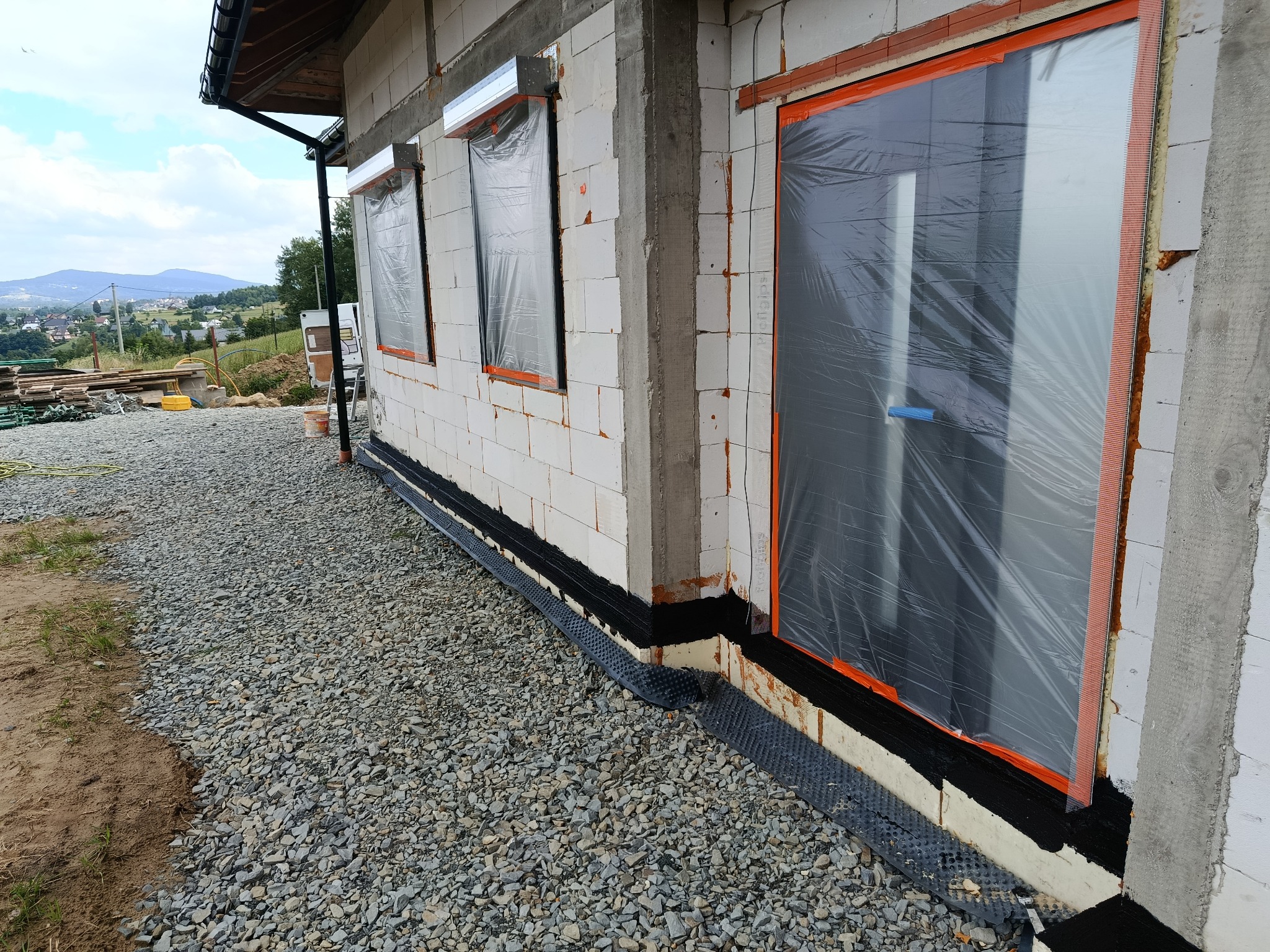 Ściana domu w trakcie budowy z oknami zabezpieczonymi folią, widoczne bloczki z betonu komórkowego, izolacja fundamentów i podsypka żwirowa wokół budynku.