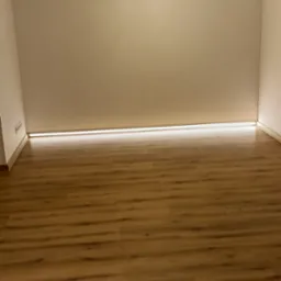 Pusty pokój z panelami podłogowymi w odcieniach brązu, oświetlony dwoma lampami sufitowymi o różnym designie i listwą LED przy podłodze.