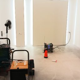 Jacek Komadowski - Przygotowanie do prac wykończeniowych w pustym, białym pomieszczeniu z podświetleniem LED, widoczny osuszacz powietrza Master, wentylator i narzędzia.