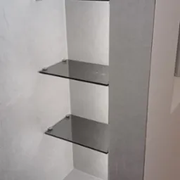 Wnęka w łazience wykończona szarymi płytkami z trzema szklanymi półkami, zamontowanymi na metalowych uchwytach.