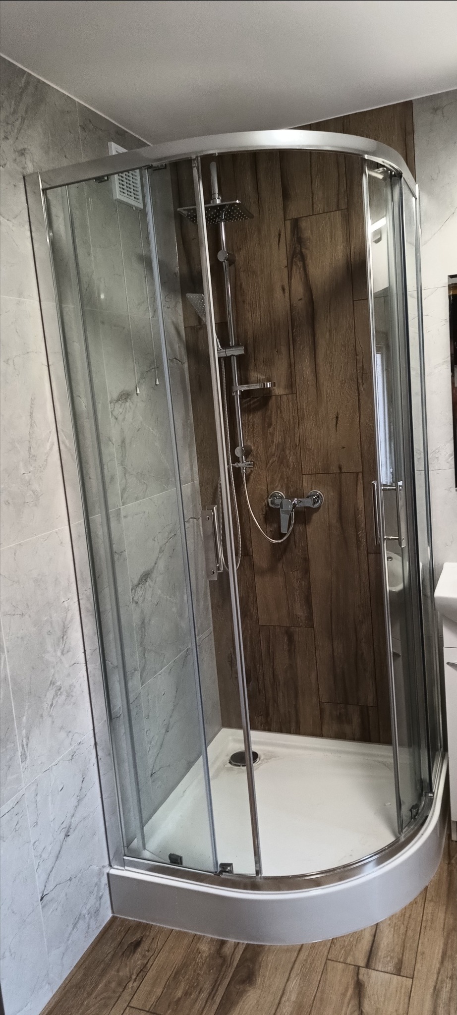 Nowoczesna kabina prysznicowa z chromowanymi elementami i panelami imitującymi drewno, zamontowana w łazience wykończonej płytkami o wzorze marmuru.