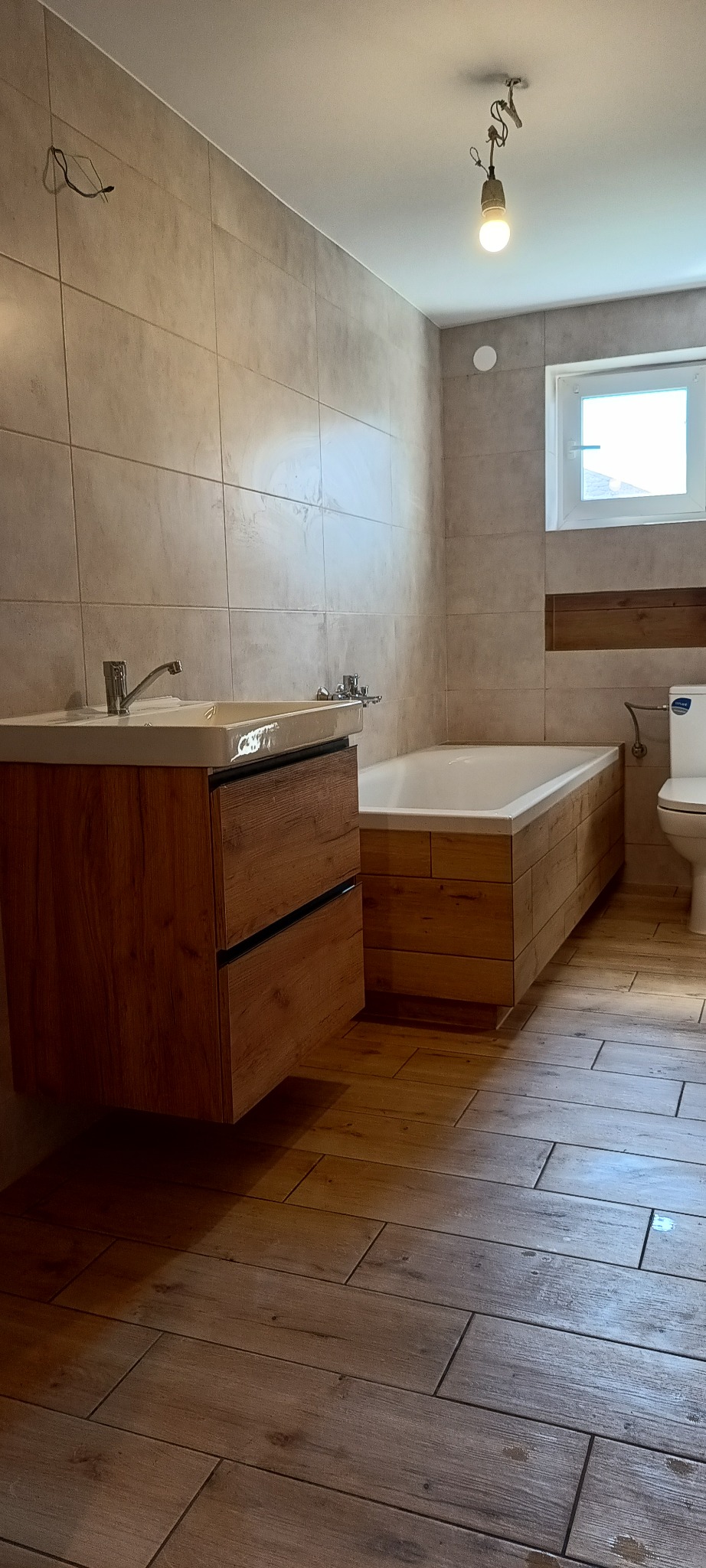 Wykończona łazienka z drewnopodobnymi płytkami na podłodze i ścianach, wisząca szafka z umywalką z blatem, obudowana wanna i toaleta w tle.