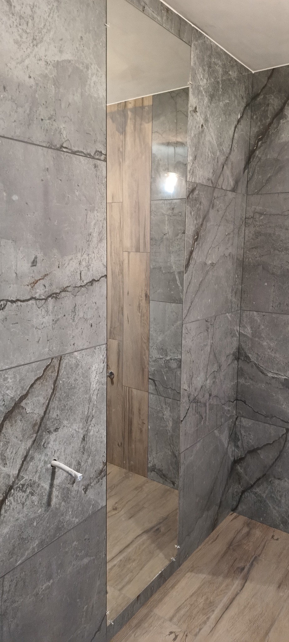 Szare płytki imitujące kamień na ścianach łazienki, z widocznym fragmentem drewnopodobnej podłogi i wystającym fragmentem rury, odbijającym się w lustrze.