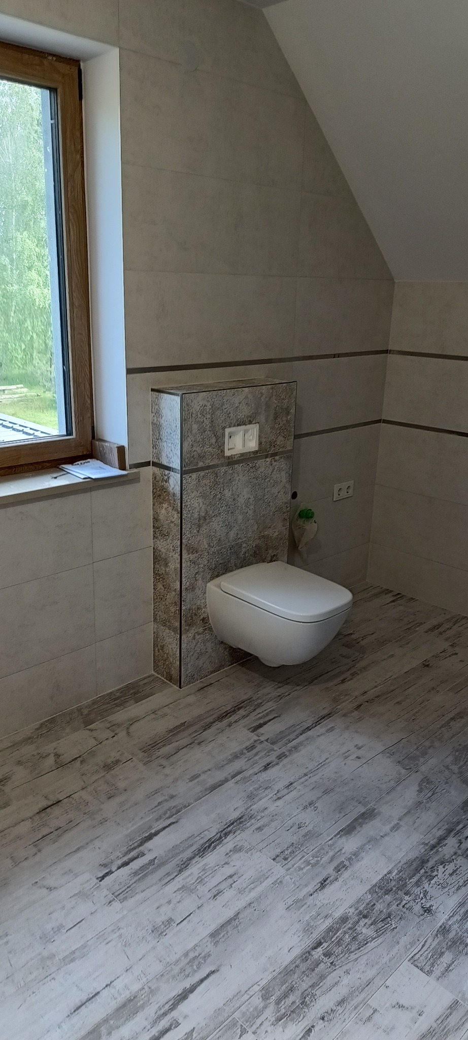 Nowoczesna łazienka na poddaszu z podwieszaną toaletą, płytkami imitującymi drewno na podłodze i beżowymi płytkami na ścianach, oświetlona naturalnym światłem z okna.