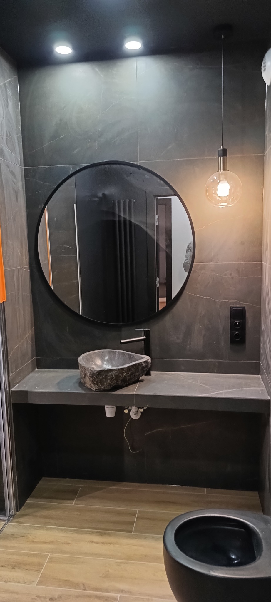 Nowoczesna łazienka z ciemnymi płytkami imitującymi marmur, kamienna umywalka nablatowa o nieregularnym kształcie, okrągłe lustro w czarnej ramie, wisząca lampa z żarówką Edisona i czarna toaleta.