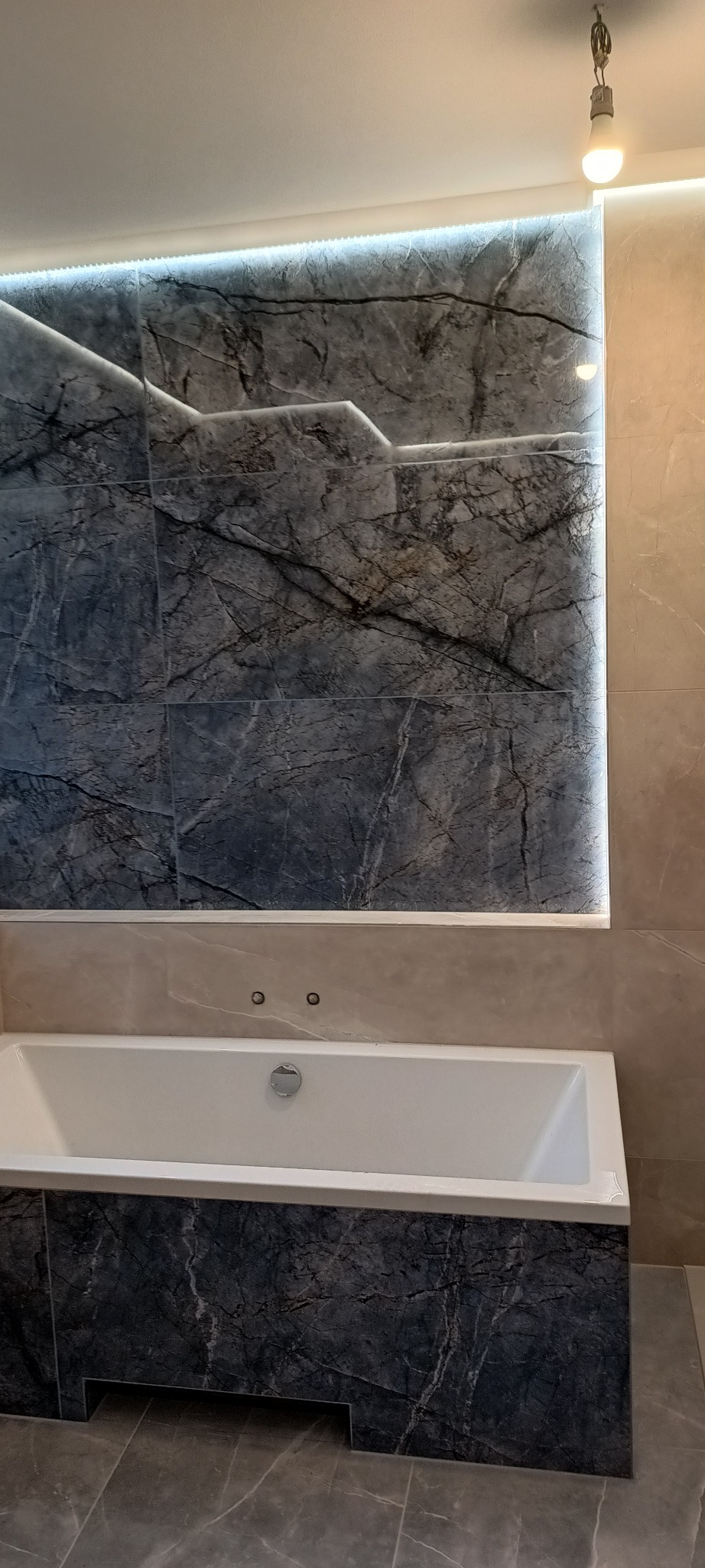 Nowoczesna łazienka z wanną obudowaną płytkami imitującymi marmur, podświetlona listwą LED na ścianie z płytek w tym samym stylu.