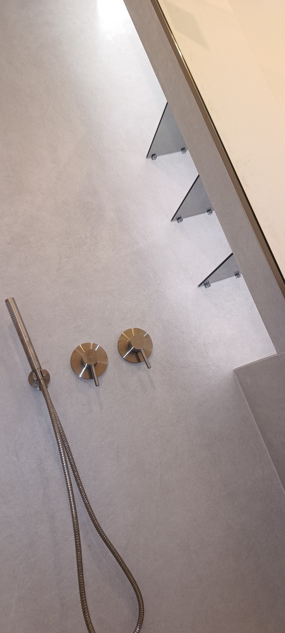 Minimalistyczny prysznic z szarą, teksturowaną ścianą, metalowymi bateriami i deszczownicą zamontowaną na szklanych półkach.