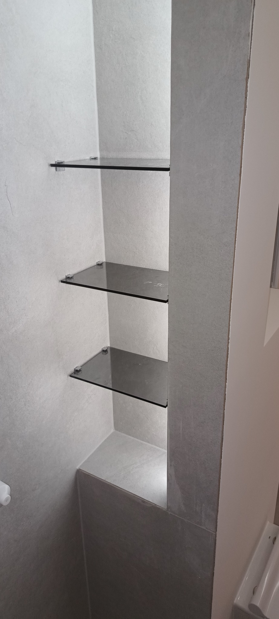 Wnęka w łazience wykończona szarymi płytkami z trzema szklanymi półkami, zamontowanymi na metalowych uchwytach.