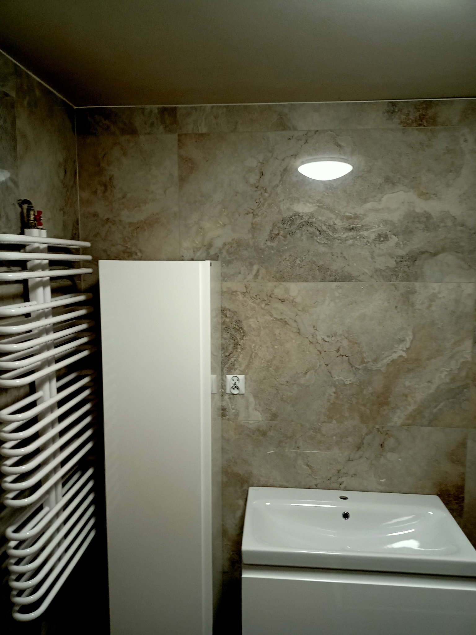 Umywalka z szafką, biały grzejnik drabinkowy i wysoka szafka w łazience wykończonej płytkami imitującymi marmur.