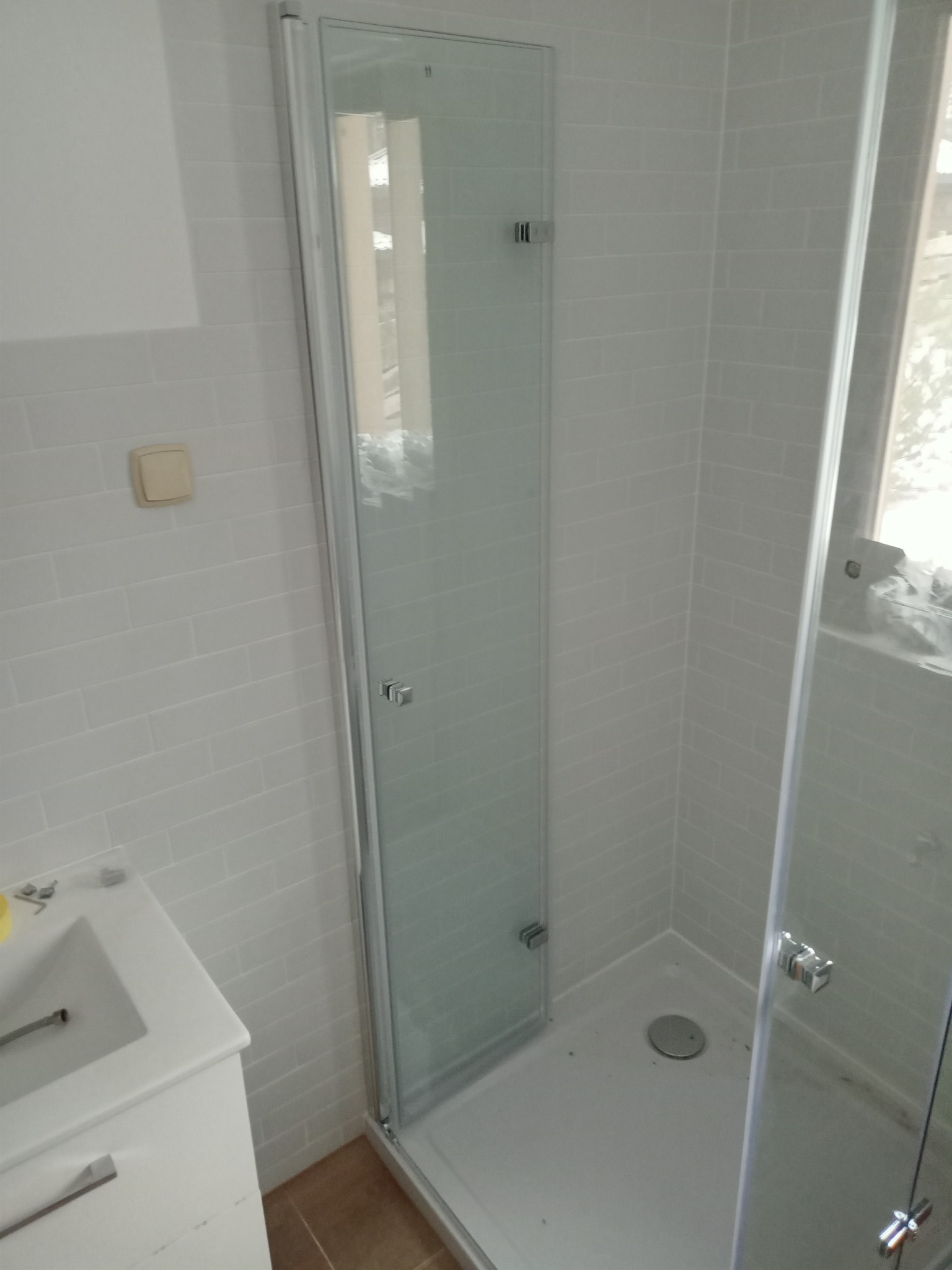 Zamknięta kabina prysznicowa z przezroczystego szkła, zamontowana w narożniku łazienki wyłożonej białymi, drobnymi płytkami, widoczna umywalka z szafką.