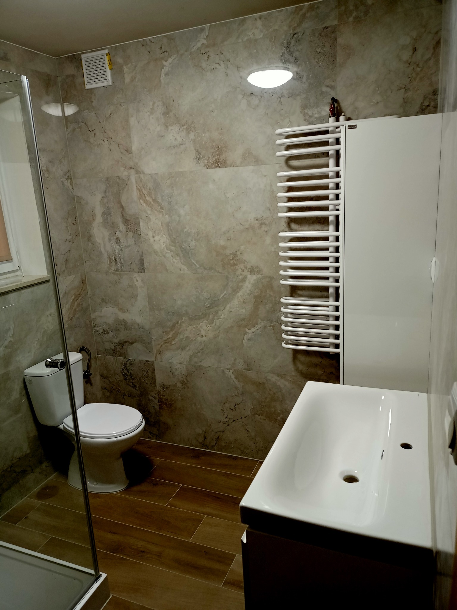 Nowoczesna łazienka z marmurowymi płytkami, drewnopodobną podłogą, białą umywalką, grzejnikiem drabinkowym i kabiną prysznicową z widocznym fragmentem szyby.