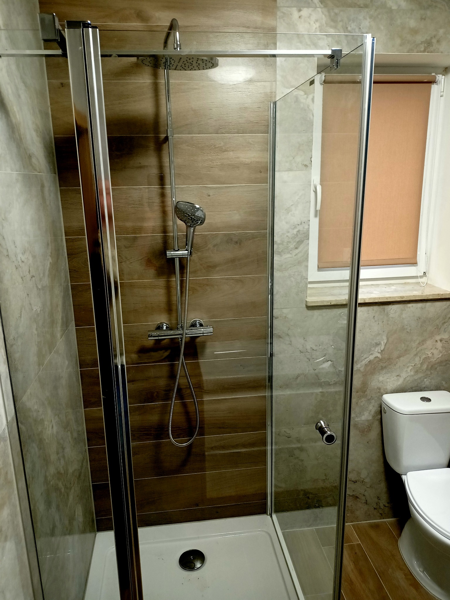 Kabina prysznicowa z chromowanymi elementami, zamontowana w łazience w Brzezinach, wykończonej płytkami imitującymi drewno i marmur, z widoczną toaletą i roletą w oknie.