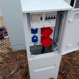 Bezpieczne Instalacje Elektryczne - Tymczasowa rozdzielnia budowlana z gniazdami w Przegini. Biała skrzynka z zabezpieczeniami i gniazdami siłowymi, widoczne przewody i uziemienie. Zasilanie placu budowy.