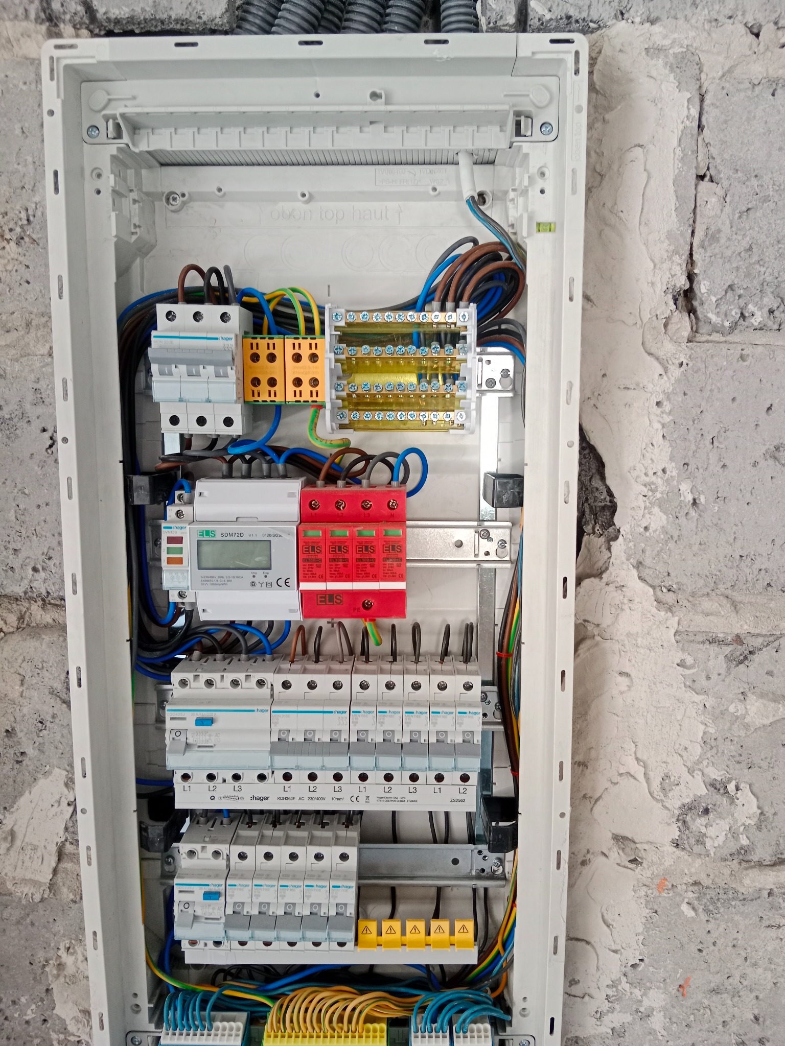 Zamknięta skrzynka elektryczna Hager z zainstalowanymi bezpiecznikami, licznikiem i przewodami, zamontowana na surowej ścianie. Widoczne kable w peszlach.