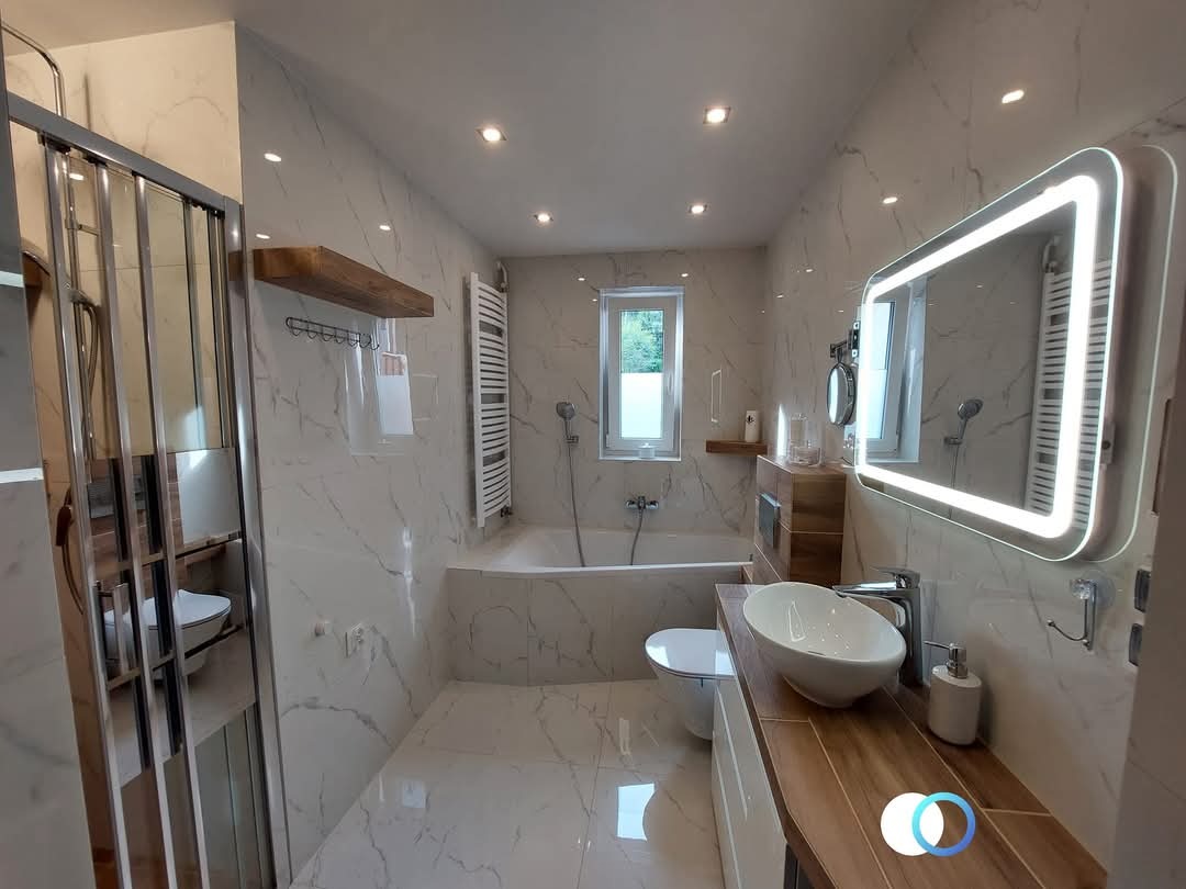 Nowoczesna łazienka z marmurowymi płytkami, podświetlanym lustrem i drewnianymi akcentami. Kabina prysznicowa ze srebrnymi ramami. Minimalistyczny design.