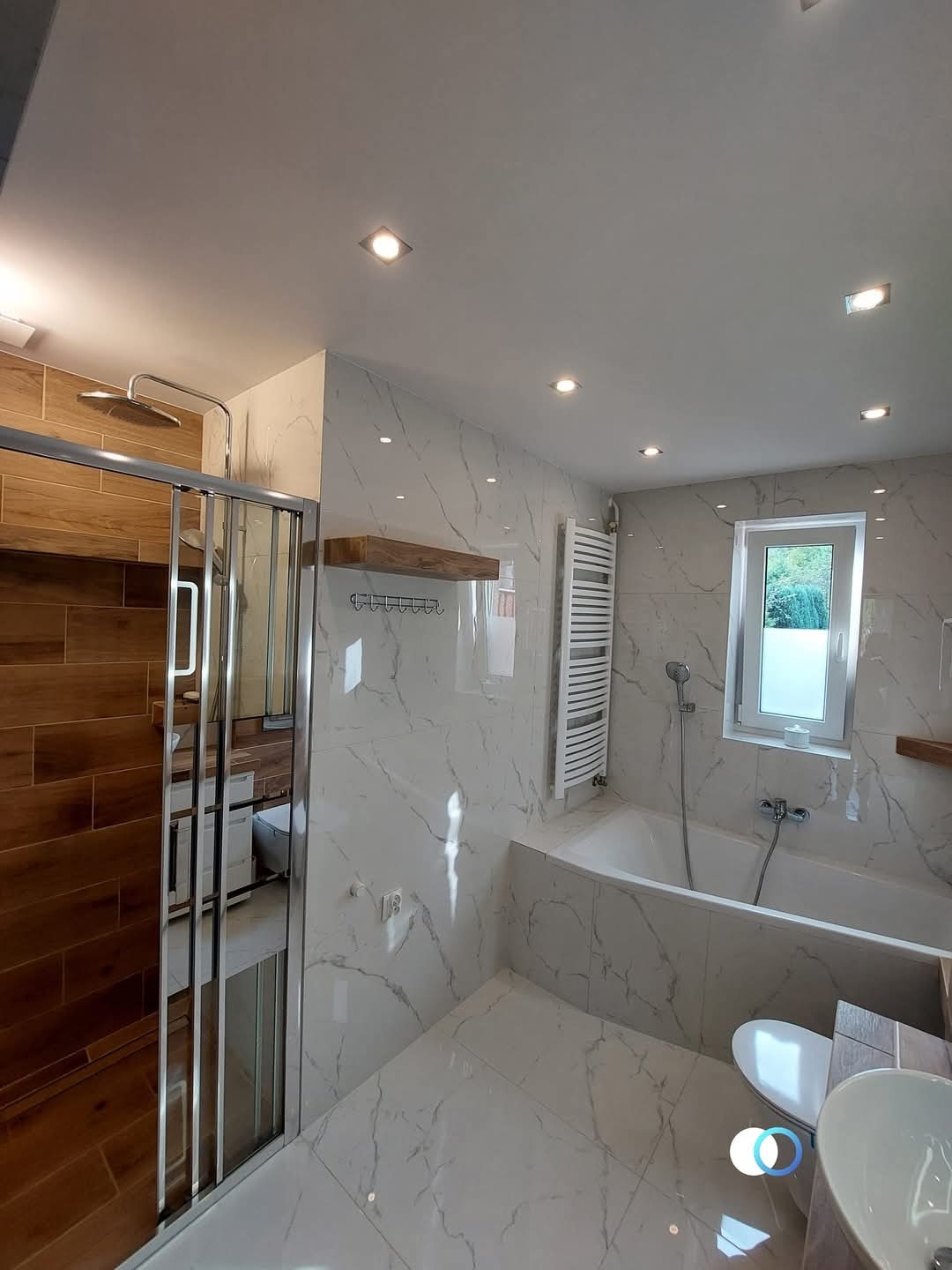 Biała łazienka z marmurowymi płytkami, kabiną prysznicową z drewnianymi panelami, grzejnikiem drabinkowym i oświetleniem punktowym w suficie.