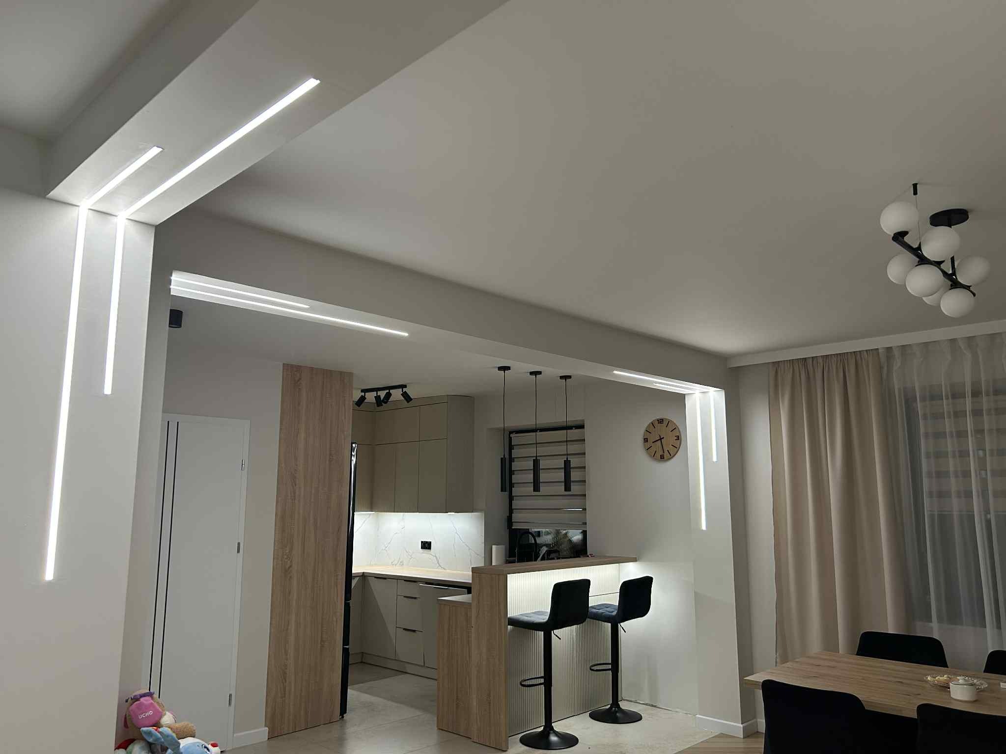 Nowoczesne oświetlenie LED w salonie z aneksem kuchennym. Listwy LED w suficie i pionowe na ścianach tworzą minimalistyczny design. Widoczny fragment kuchni z hokerami i zegar.