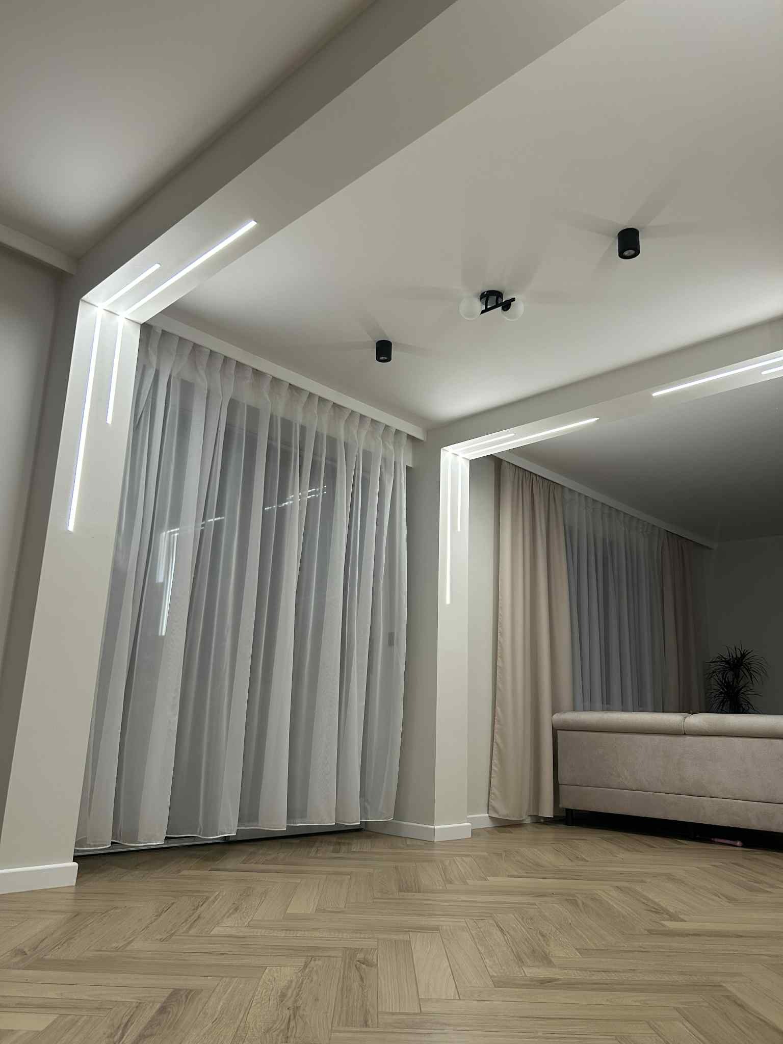 Nowoczesne oświetlenie LED w salonie z jasnymi zasłonami i parkietem w jodełkę. Czarny reflektor sufitowy dodaje kontrastu.