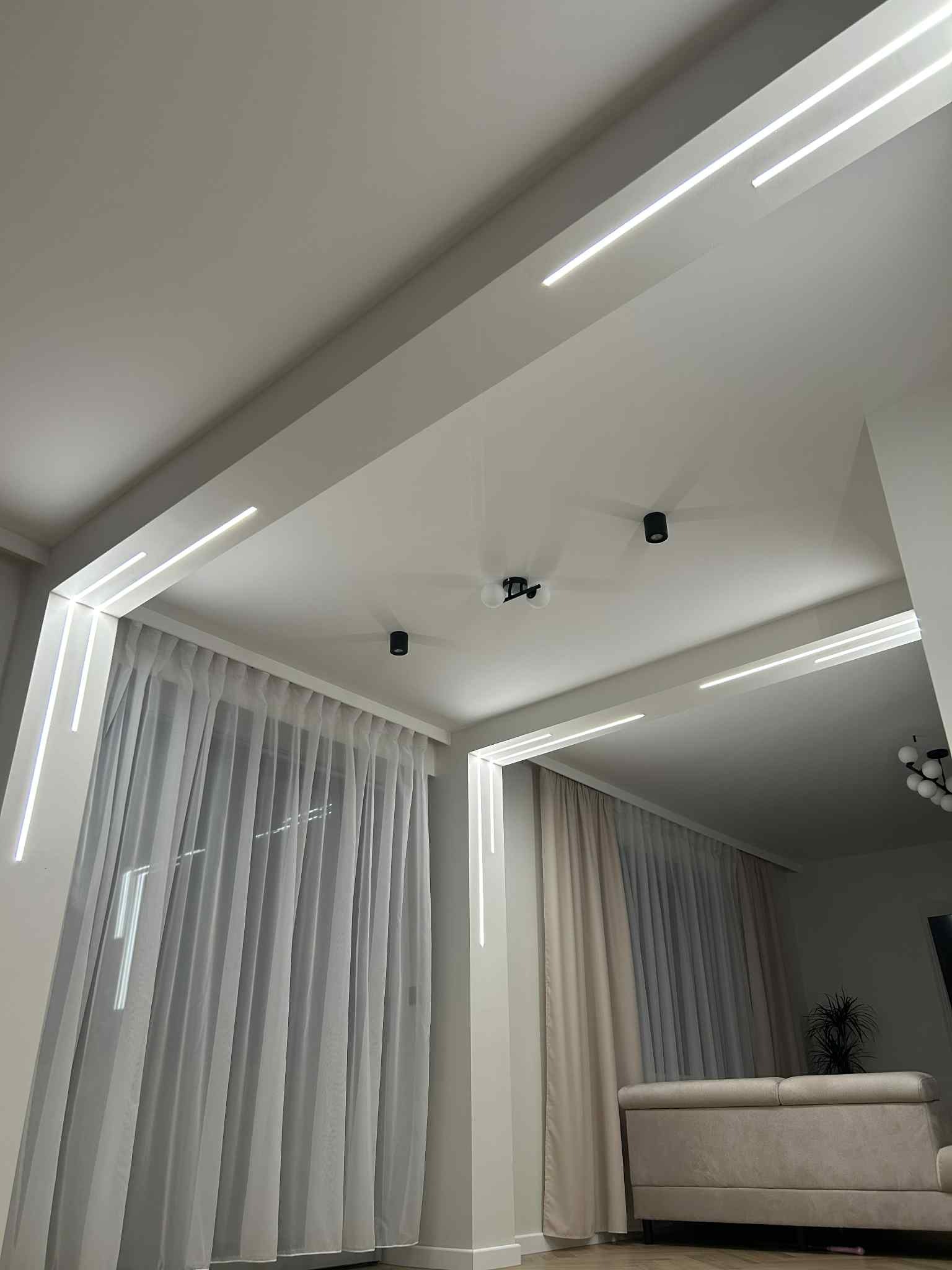 Nowoczesne oświetlenie LED w suficie podwieszanym, zintegrowane z karniszami i punktowymi lampami sufitowymi w jasnym salonie z zasłonami.